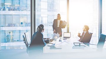 Cheerful female CEO in formal suit standing near table with laptop and explaining documents to group of coworkers during meeting in modern office