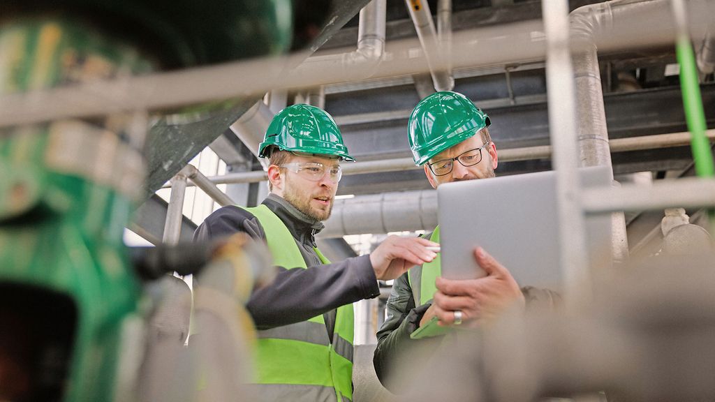 Low angle view of manual workers discussing while using laptop. They are standing amidst machinery. Professionals are in protective workwear.