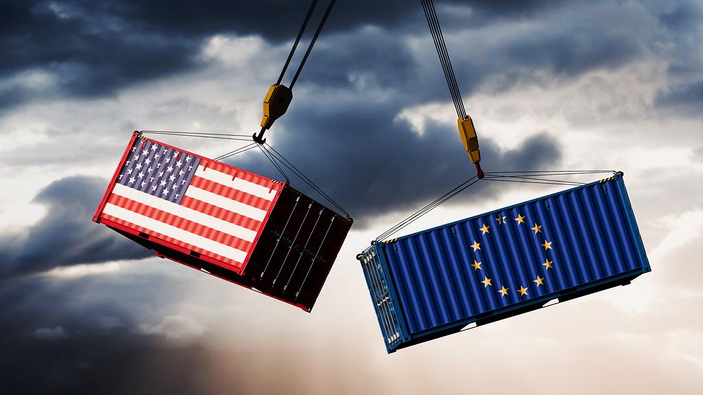 Zwei Container mit den Flaggen der USA und der EU baumeln an einem Kran in der Luft. 