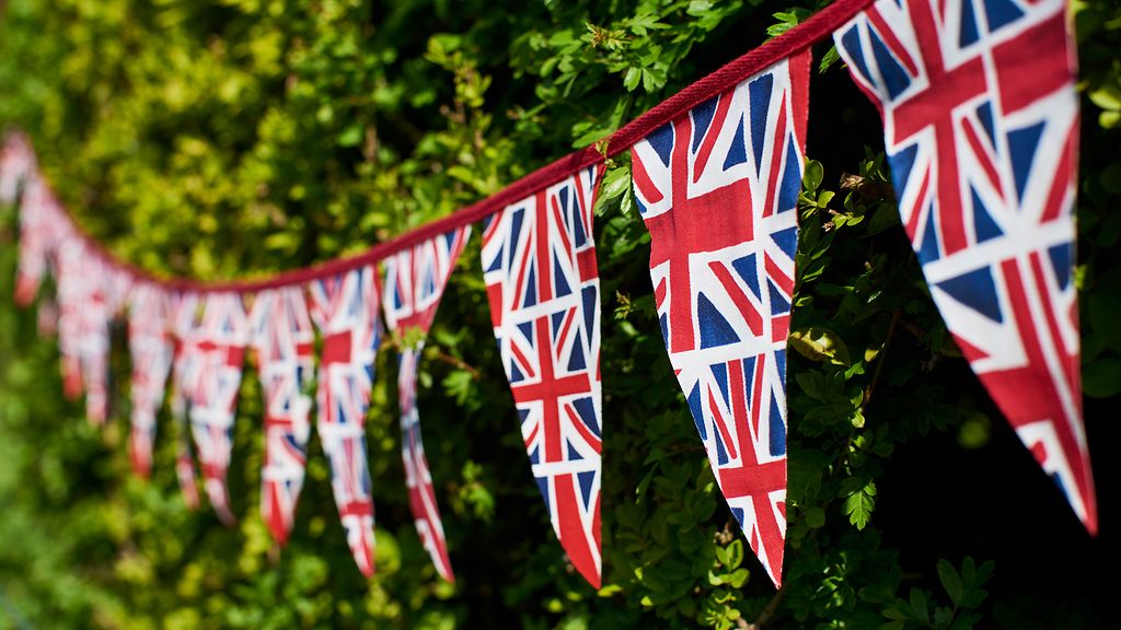 Wimpelkette mit britischen Flaggen vor einer grünen Hecke. 