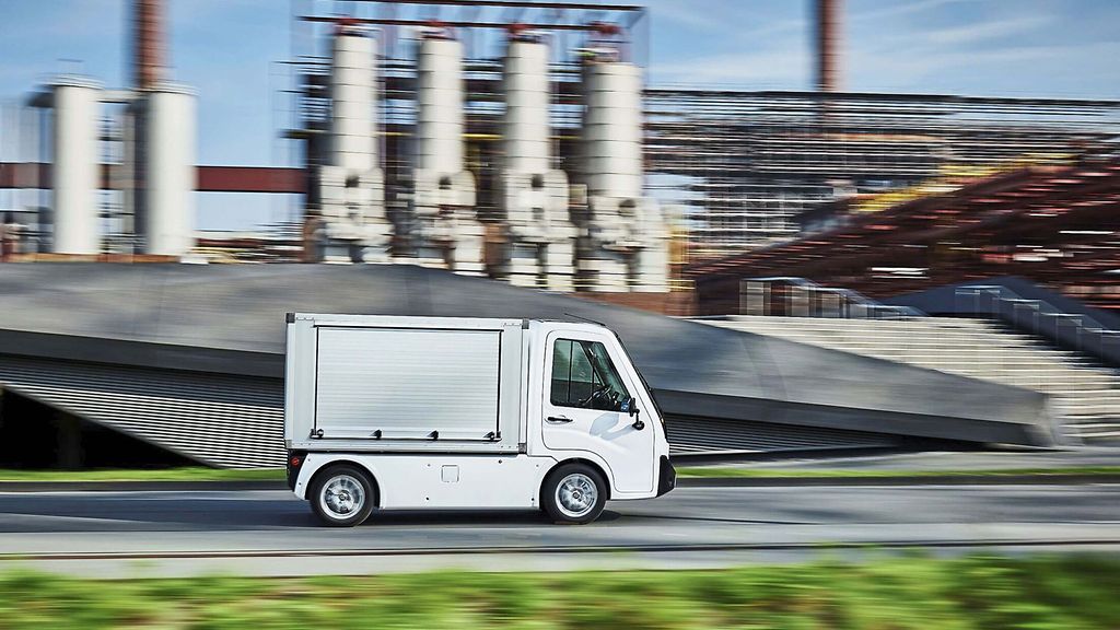 Kleiner weißer Elektrotransporter fährt seitlich im Profil an einer Industrieanlage vorbei. Unscharfer Hintergrund betont die Bewegung des Fahrzeugs.