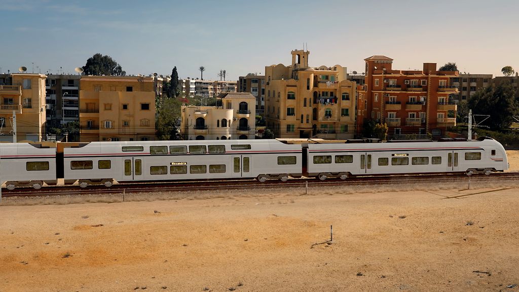 Ein ICE-Schnellzug fährt auf einer Bahnstrecke am Rand eines Wohnviertels in wüstenähnlicher Umgebung durch Ägypten.