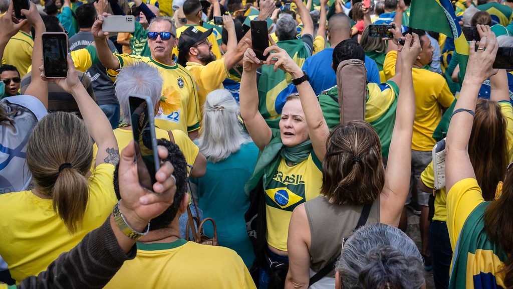 Dicht gedrängte Menschenmenge in gelber und grüner Kleidung mit Brasilienflaggen, die ihre Smartphones hochhält und feiert. 