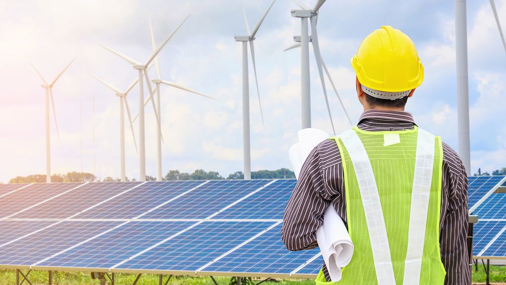 Junger Kaufmann Ingenieur mit gelbem Helm am Solarmodul und Windgeneratoren Kraftwerksbaustelle Hintergrund