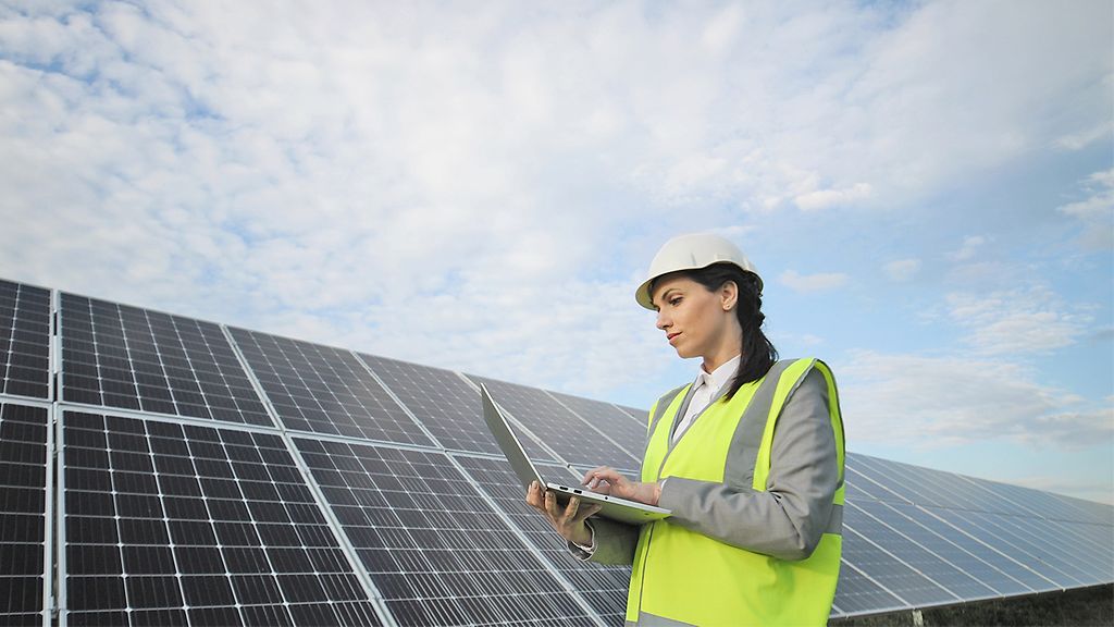 Portrait of electrician engineer in safety helmet and uniform using laptop checking solar panels. Female technician at solar station.