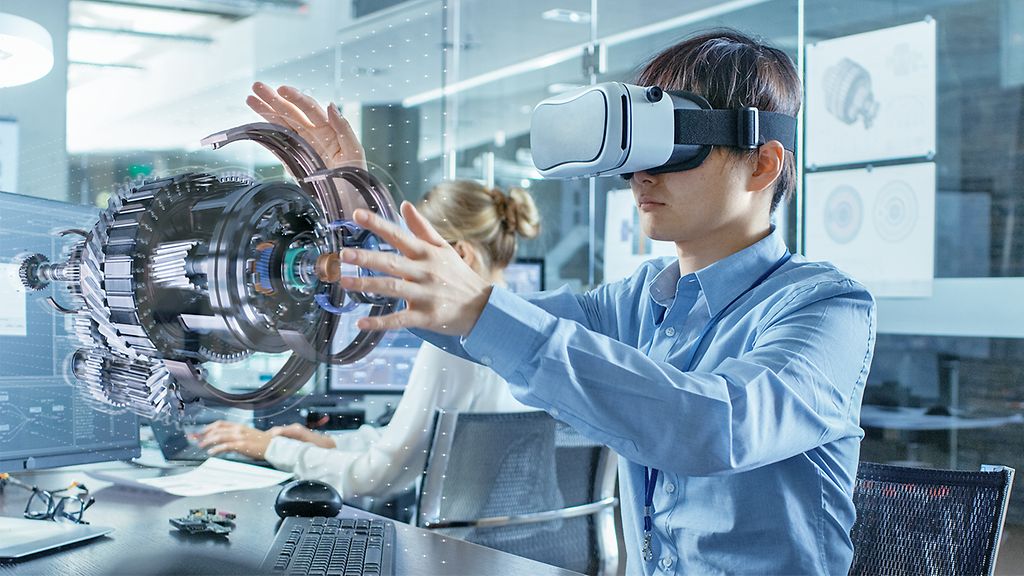 Computer Science Engineer wearing Virtual Reality Headset Works with 3D Model Hologram Visualization, Makes Gestures. In the Background Engineering Bureau with Busy Coworkers.