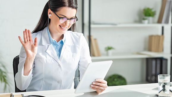 positive doctor waving and having online consultation on digital tablet in clinic office with laptop