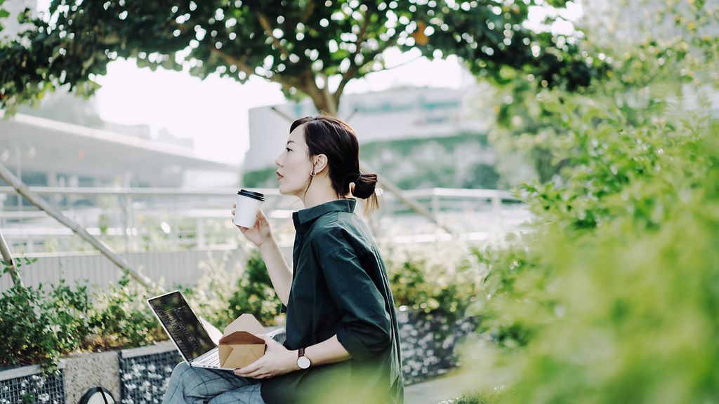 Professional young Asian businesswoman sitting on the bench in an urban park, working on laptop while having a healthy salad lunch box with a cup of coffee during lunch break, asiatische Geschäftsfrau, Mittagspause, Stadtpark