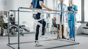 Modern Hospital Physical Therapy: Patient with Injury Walks Wearing Advanced Robotic Exoskeleton. Physiotherapy Rehabilitation Scientists, Engineers use Tablet Computer to Help Disabled Person, modernes Krankenhaus, Physiotherapie, Patient, Roboter-Exoskelett