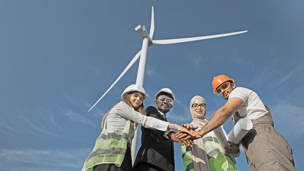Group of four multicultural partners stacking hands together and smiling on camera while standing among wind turbines. Men and women in safety helmets having successful meeting outdoors.